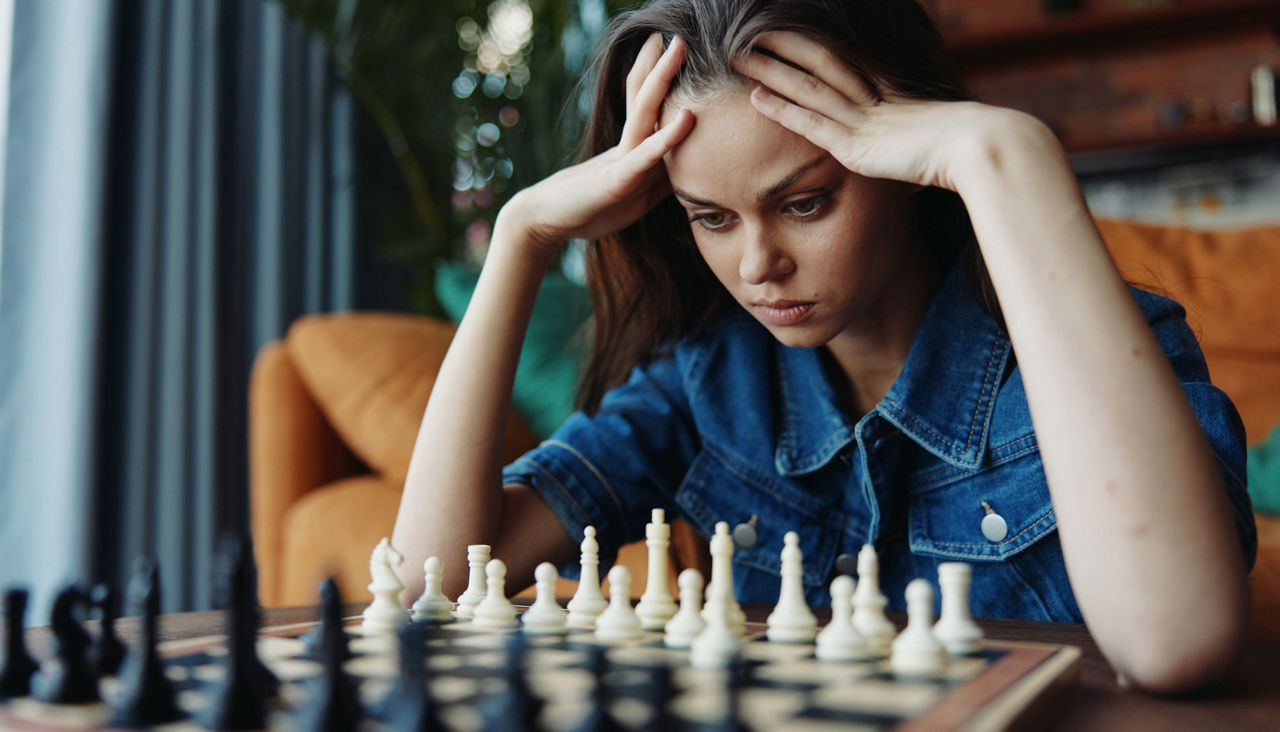 Young woman studies chess game at table, head in hands, deep in thought