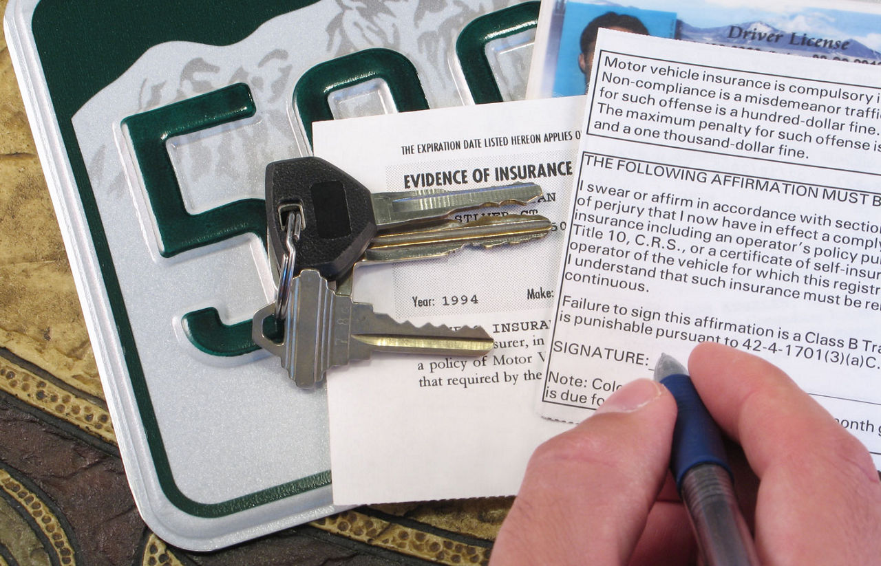 A person signing a car title with the keys, an insurance card and the license plate in the background.