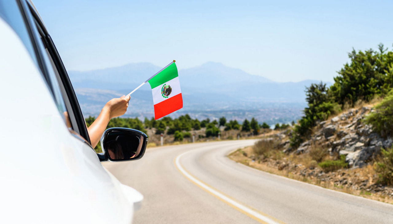 Woman holding Mexico flag from the open car window driving along the serpentine road in the mountains.