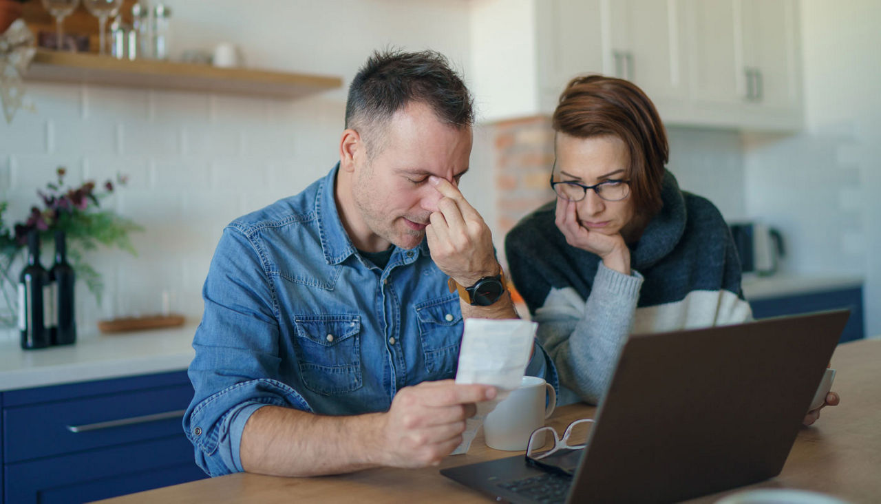 Stressed couple sitting at kitchen table with bills, checks and laptop 