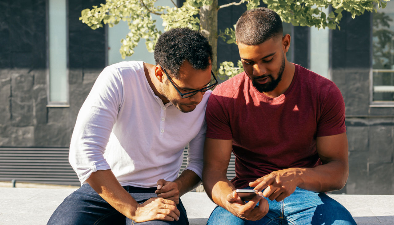 Two young adult men look with concentration at a cell phone one is holding and pointing at