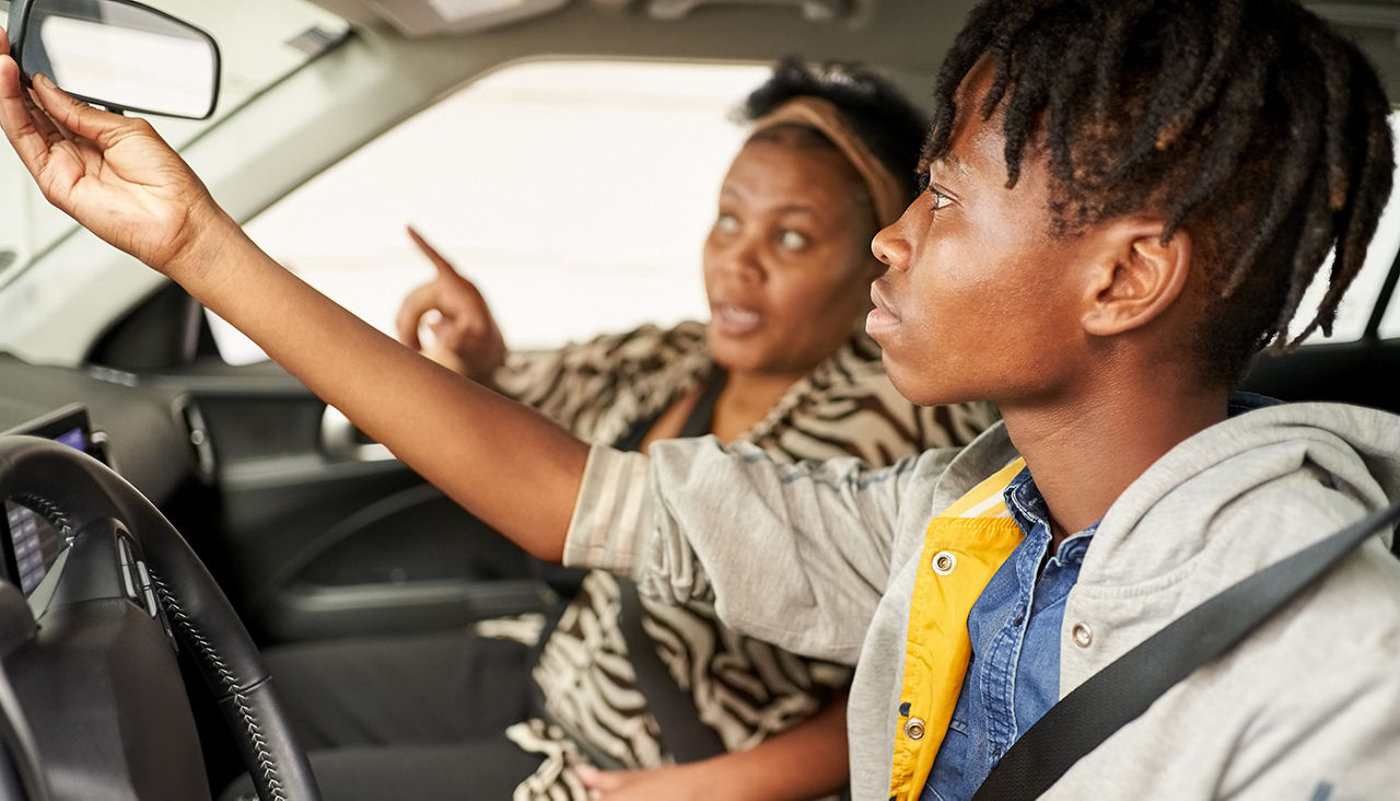 Teenage boy adjusting rear view mirror while learning to drive car with his mother