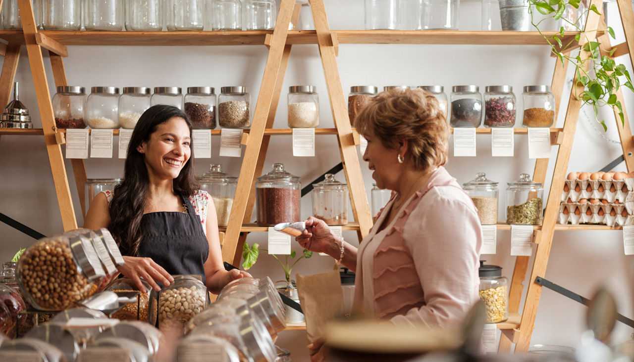 Happy hispanic employee and senior female customer standing next to the shelves of a sustainable shop and smiling at each other.