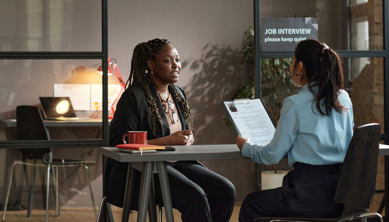 African young woman talking to manager at table at job interview while she examining her resume
