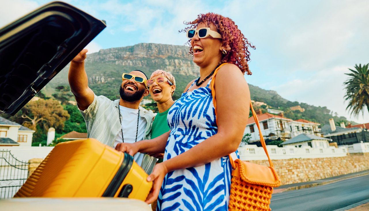 Three friends on vacation smiling and laughing as one lifts suitcase out of car trunk