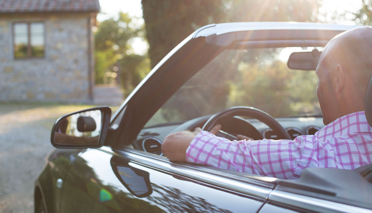 A man driving a convertible up a long driveway in the sunlight