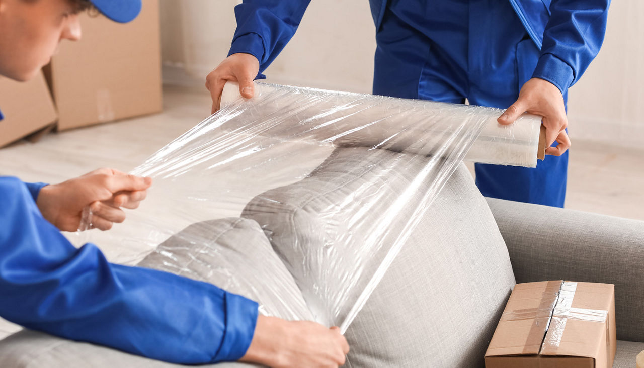 Male movers wrapping sofa with stretch film in room