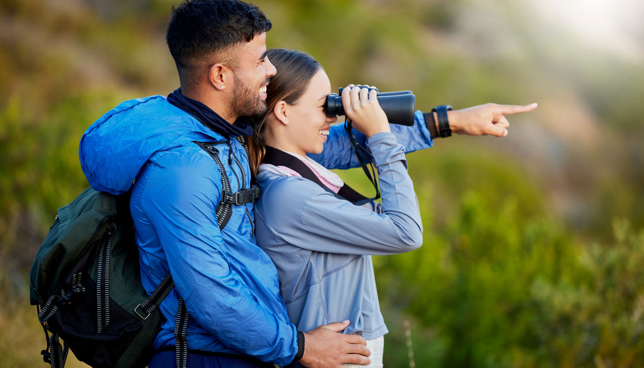 Young man and woman sightseeing in mountains smiling as woman looks through binoculars