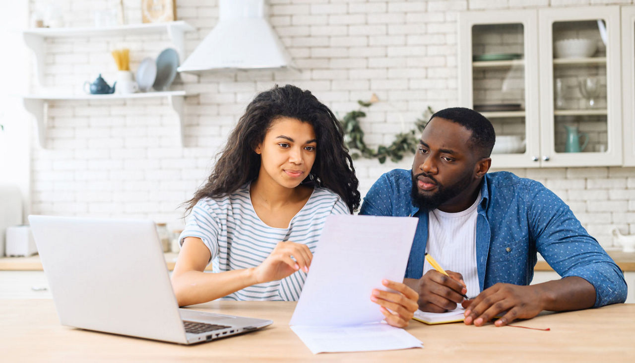 A couple sits at their kitchen table, working with documents and a laptop