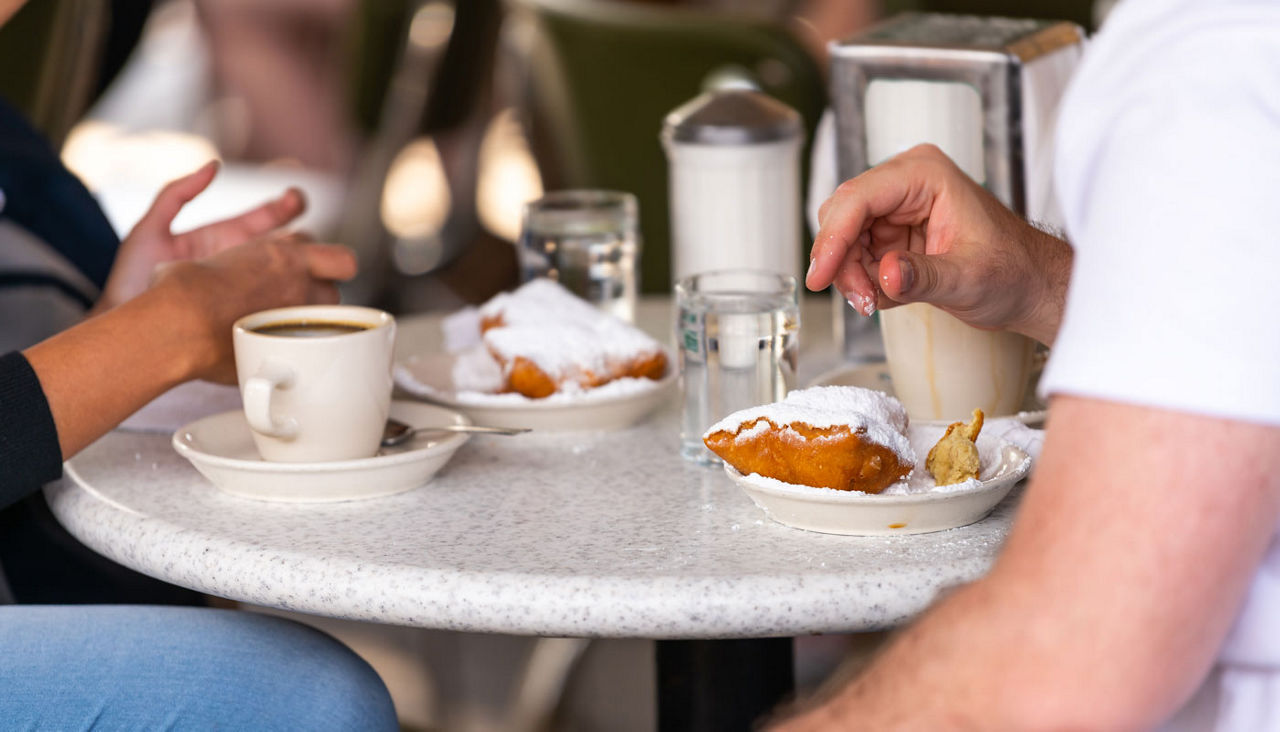 couple sitting outside at sidewalk cafe by table drinking chicory coffee and eating deep fried beignet donut powdered with sugar