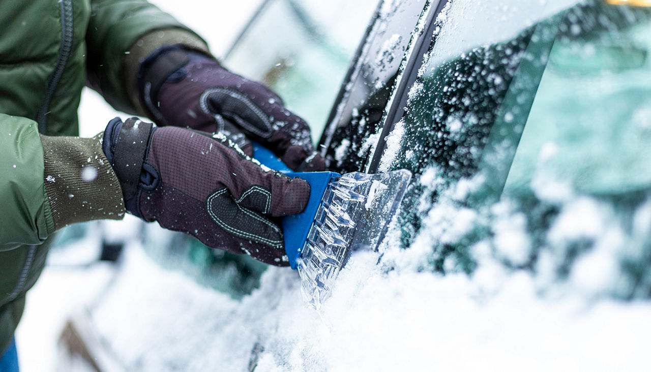 Color image depicting a senior man de-icing his snow-covered car in a blizzard in winter. The man is wearing warm winter hat and gloves and houses on a residential street are defocused in the background. Room for copy space.