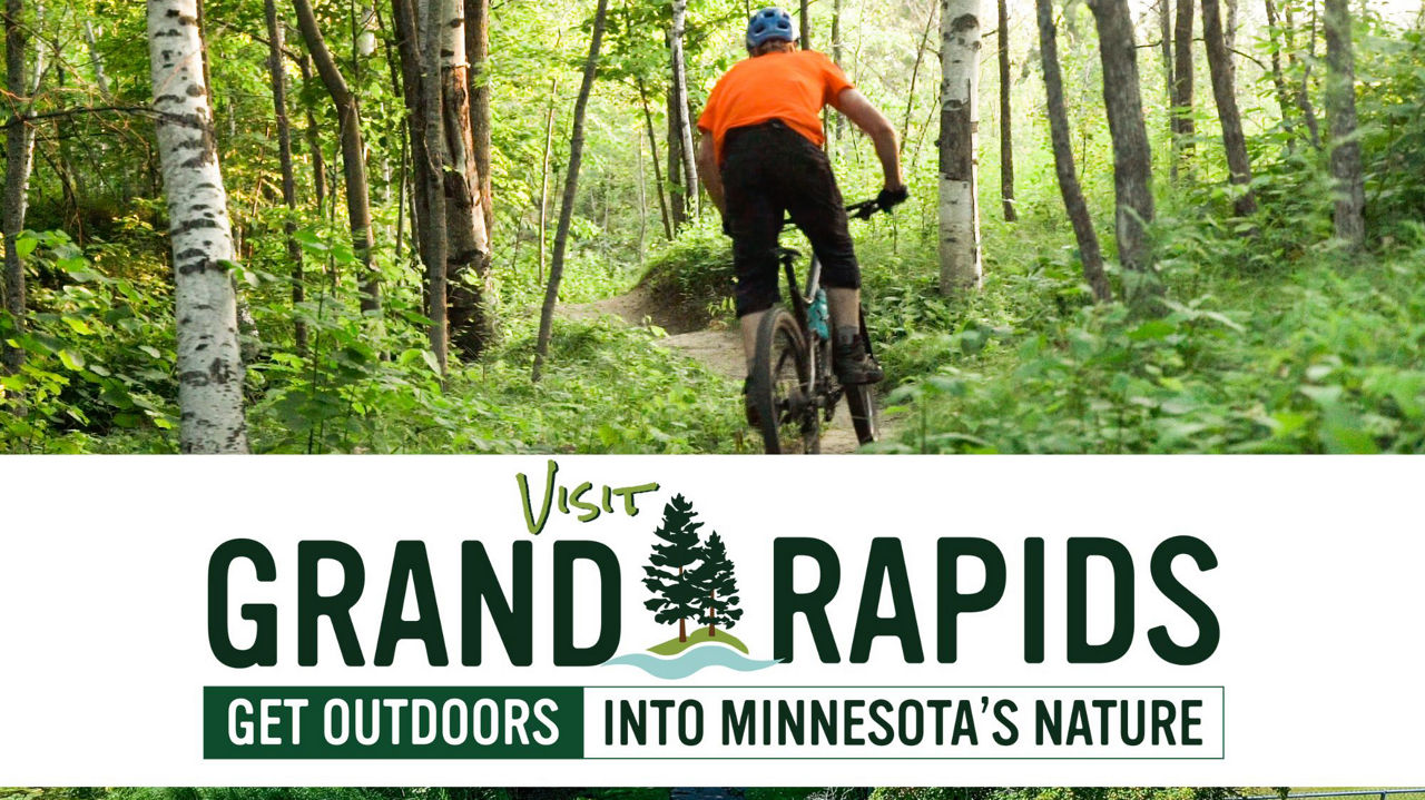 Visit Grand Rapids logo.  Get outdoors into Minnesota's nature. Photo of person in black bike shorts, orange short sleeve shirt wearing a bike helmet on a bike on a trail through trees. 