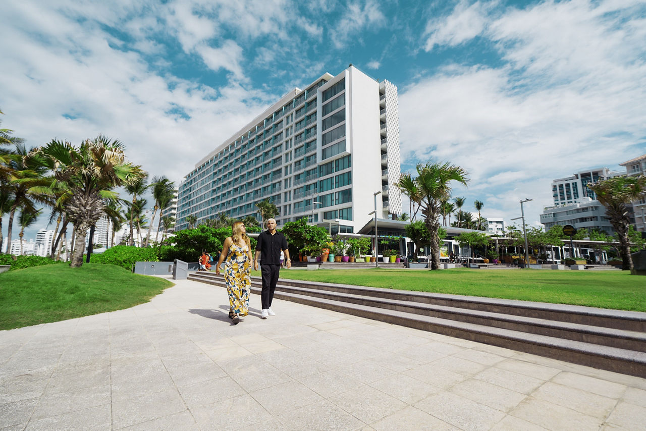 A man and a woman walking on a sidewalk in front of the La Concha Renaissance San Juan Resort. Blue sky with white puffy clouds.
