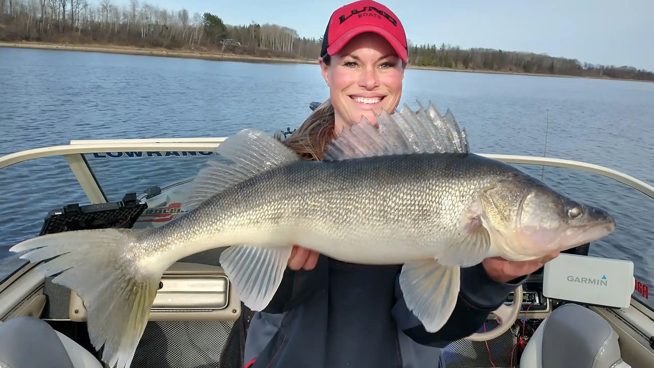 A young woman holding a large fish while standing in a boat on a lake.