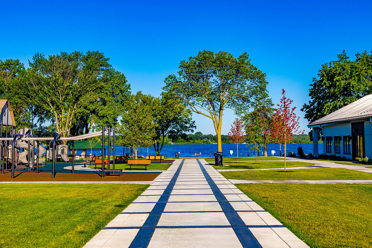 Photo of a park with playground equipment, trees and a lake. A sidewalk lined by grass leads to the water.