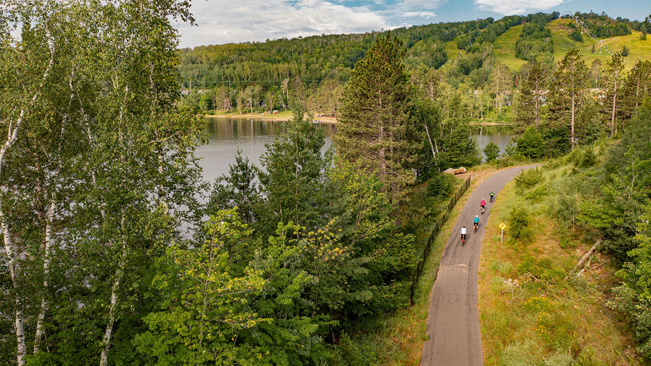 Aerial view of four bike riders on a trail lined by trees next to a lake.