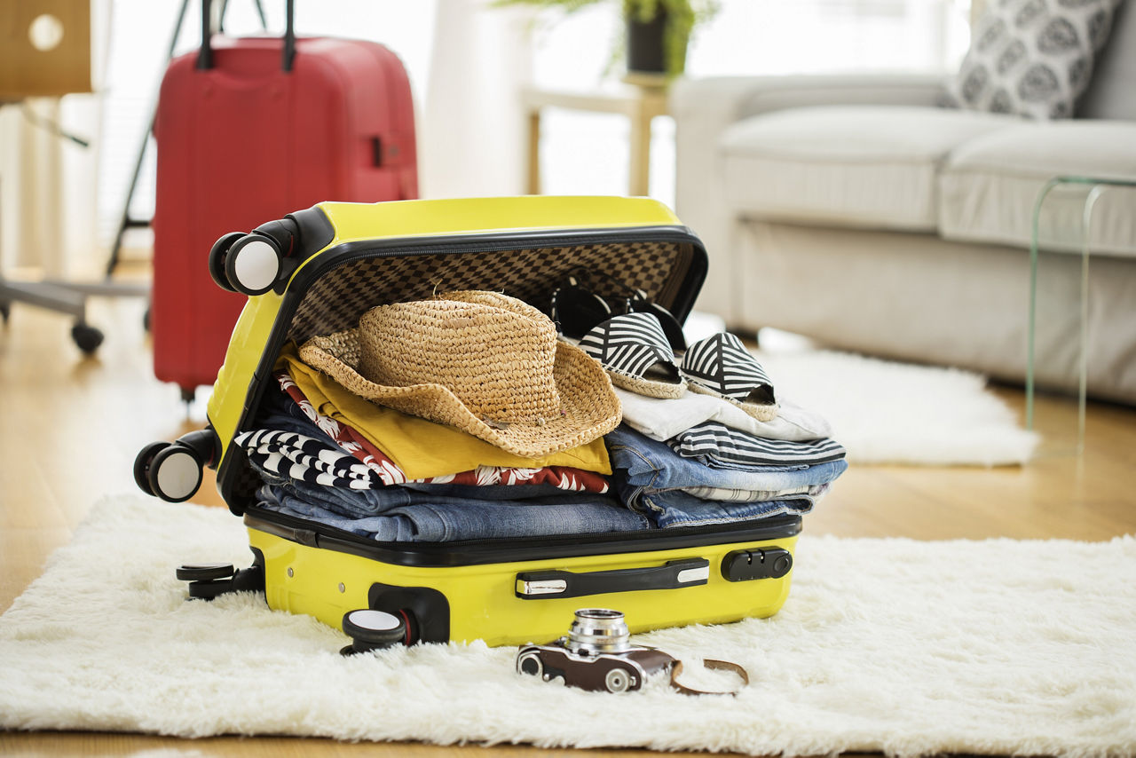An open yellow suitcase on a white, fuzzy carpet overfilled with clothing. A camera sits on the carpet in front of the suitcase. A red, closed rolling suitcase, a white couch and a wood end table are in the background.