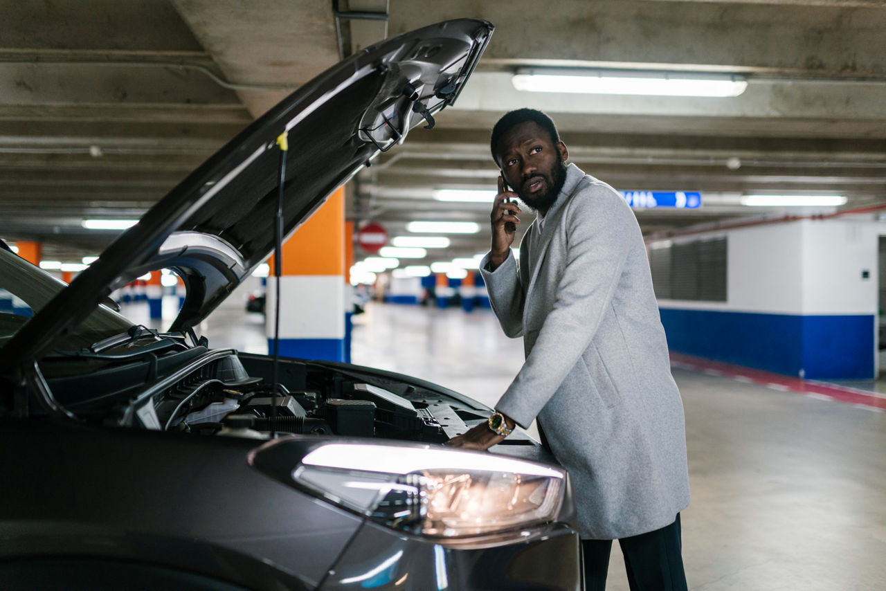 a man in front of his car with the hood up, calling someone on his phone.