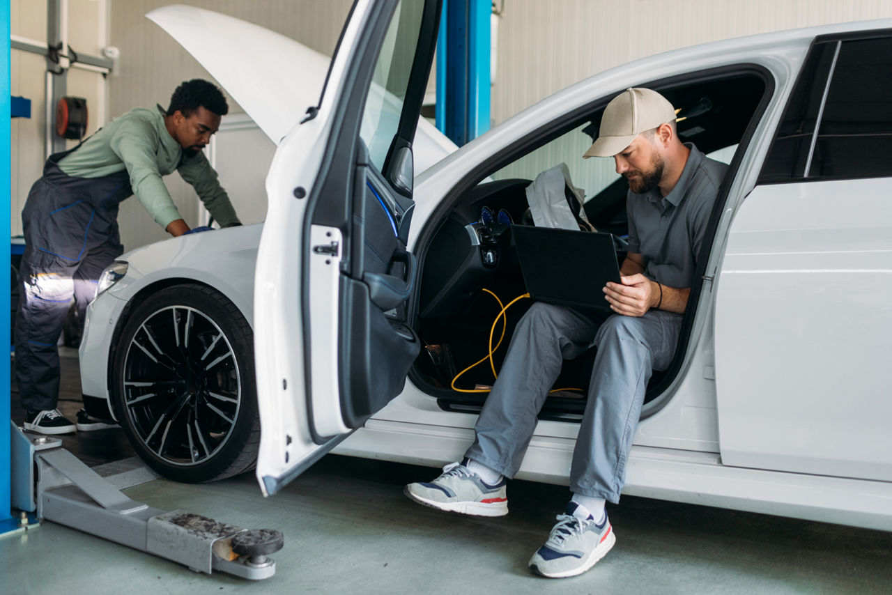 mechanic opening the hood of the car to investigate as another man is in the front seat reading.