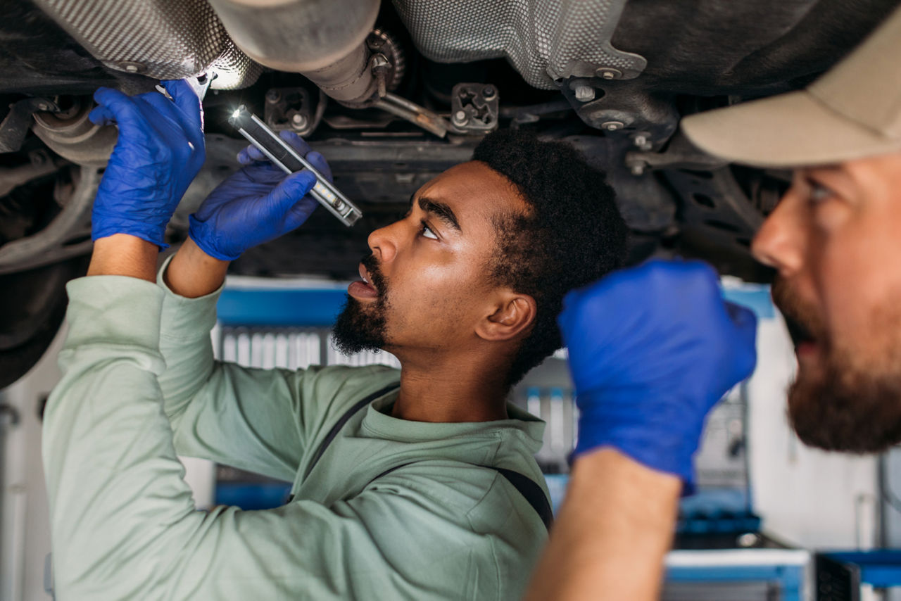 mechanic inspecting the underside of a car with another man watching him