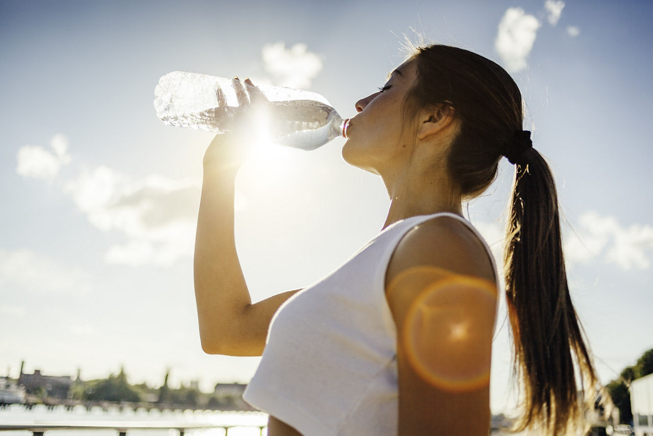 A woman with a ponytail wearing a white tank top drinking out of a water bottle.