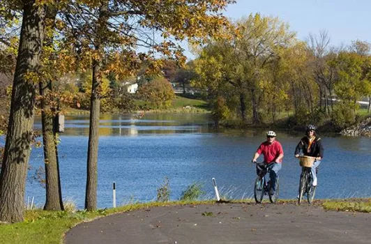 Two people on bikes on a paved trail with a lake and trees in the background.