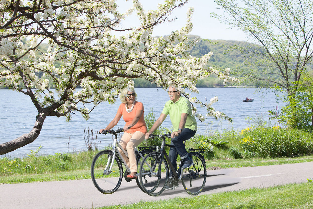 Two people wearing summer clothes on bicycles on a paved path by a lake.
