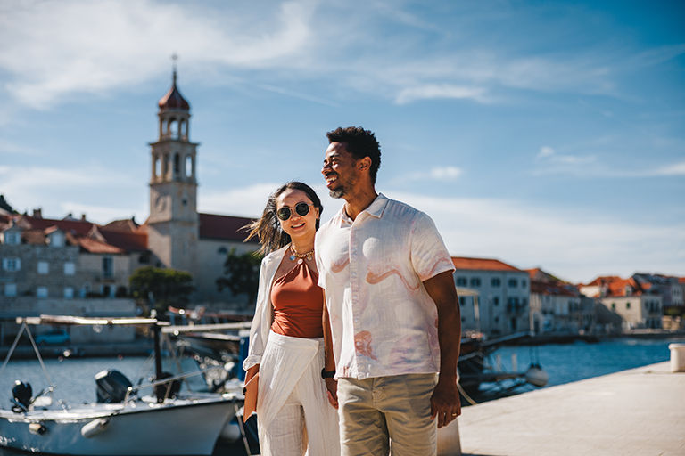 Mid adult diverse couple relishing a scenic stroll along Sutivan harbor, enjoying sunny weather and charming waterfront views. Both appear relaxed and content as they walk beside moored boats and historic architecture.
