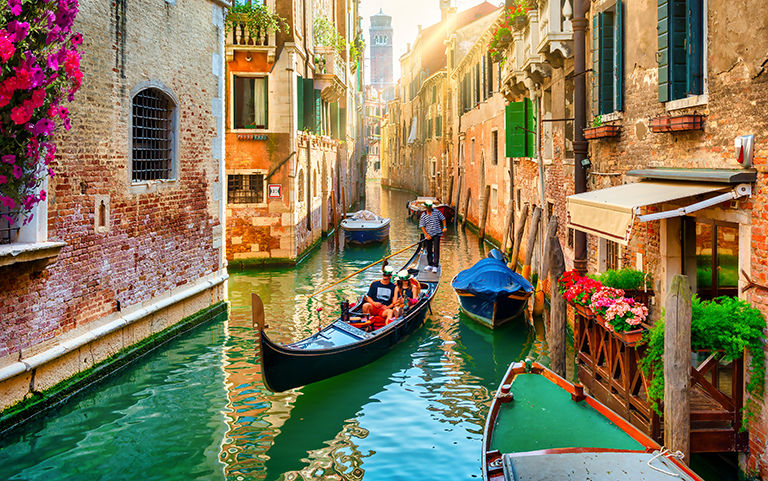 A scenic view of Venice’s historic canals, featuring traditional gondolas, colorful waterfront buildings and vibrant flowers along the waterways.