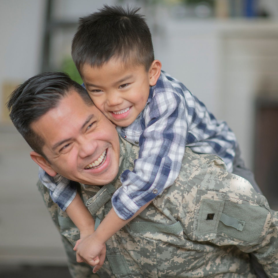A father in military uniform smiling with his son on his back smiling