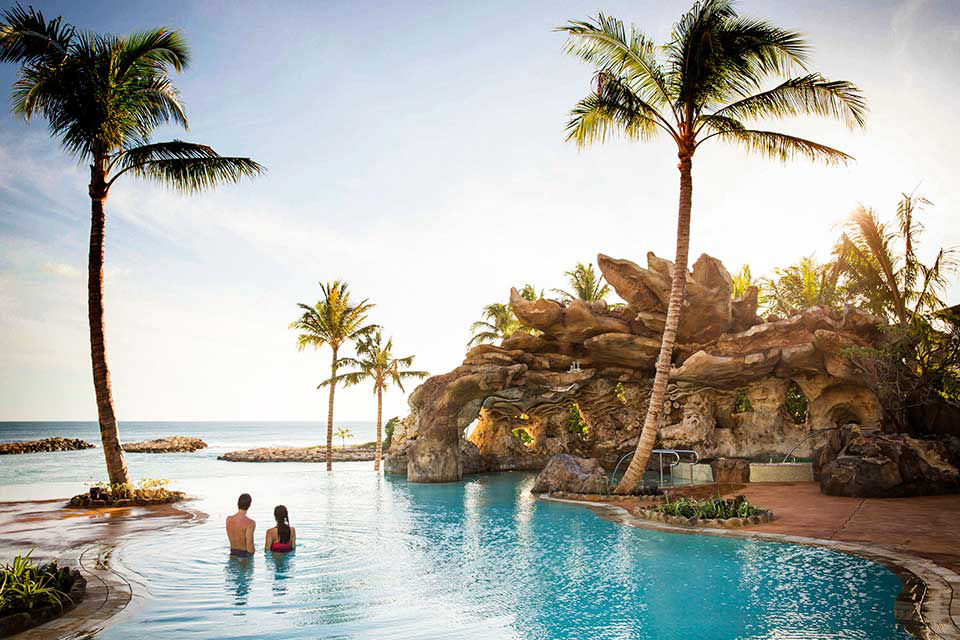 Couple swimming in pool at aulani resort facing the ocean at sunset.