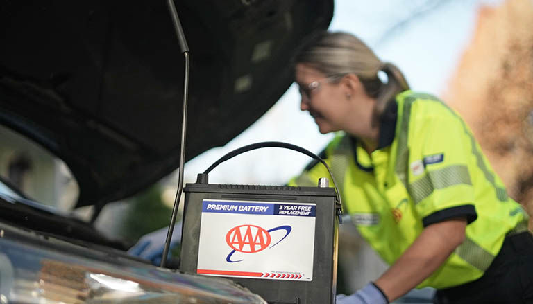 AAA technician in uniform stands beside a car, replacing the car battery. 