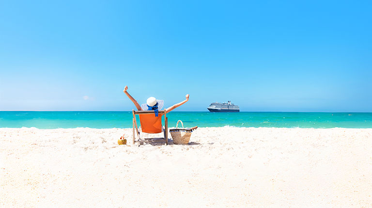A young woman sits on the shore, looking out at vibrant turquoise water and a passing cruise ship.