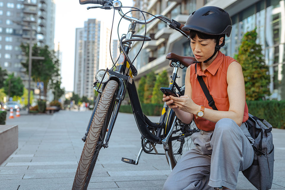 A woman wearing a helmet, crouching near her bike, looking at her phone in a city environment.