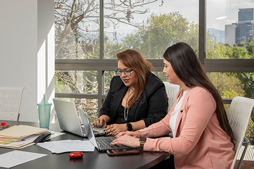 Two Latina businesswomen work focused on their laptops sitting at a desk in an office boardroom. Two women working in offices