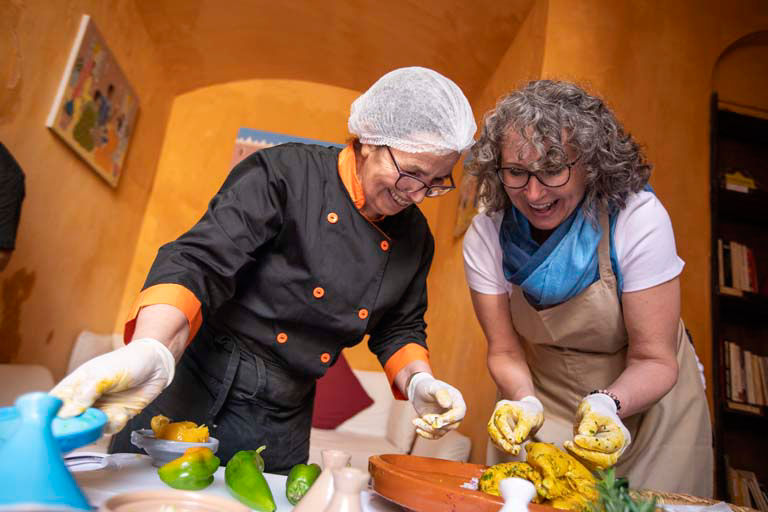 A private cooking class scene showing a chef in professional attire with hat and apron working alongside a participant in casual cooking attire