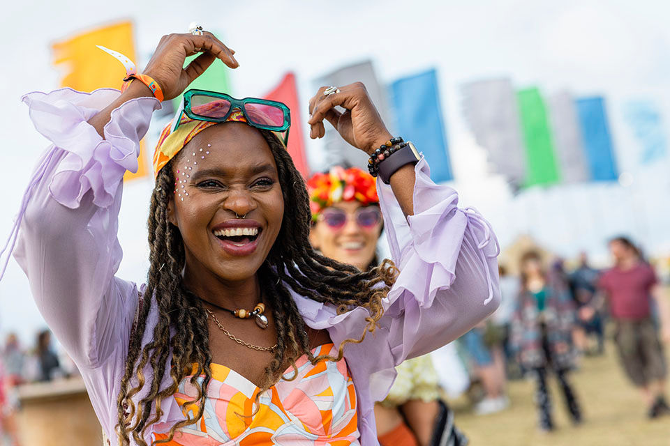 A shot of a young woman captured in the moment at a festival. She is smiling with excitement and joy. She is wearing vibrant clothing with sunglasses and a bandana.