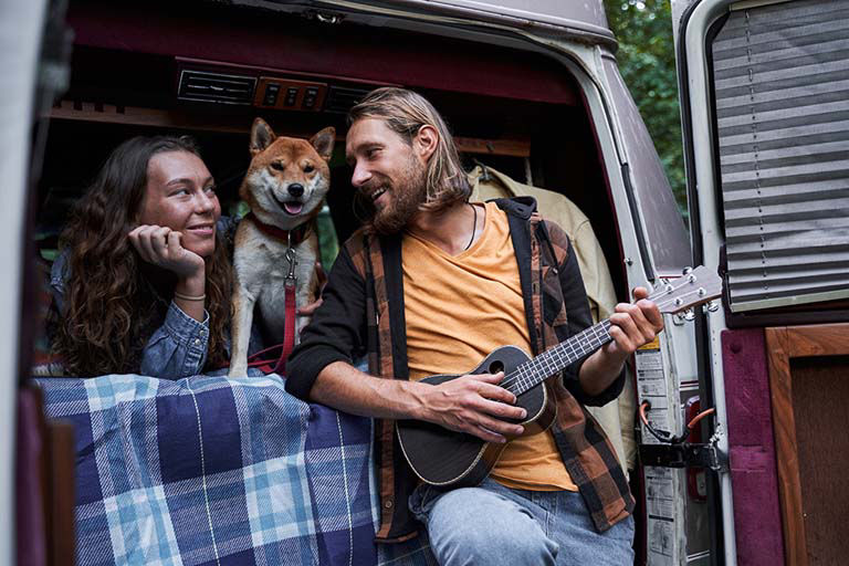 A couple on a road trip with their dog at the back of their van while he plays a small guitar.