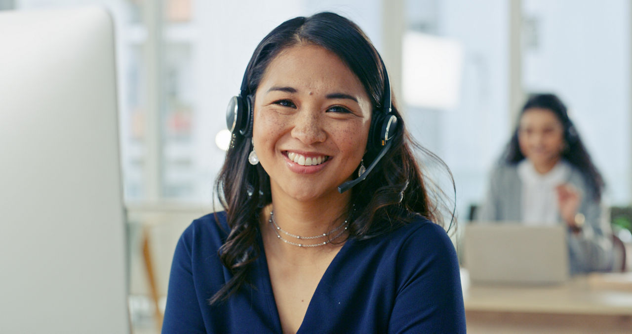 Friendly customer service representative wearing a headset in a bright office environment with colleagues in the background. The image portrays professionalism, teamwork, and a positive workplace atmosphere.