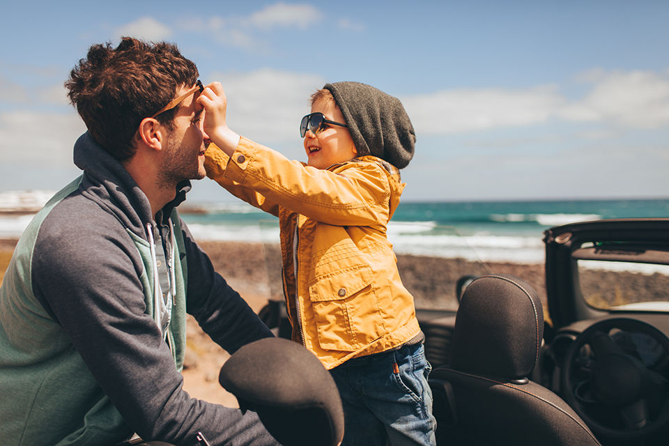 Photo of a little boy and his father on a road trip by the seaside in their convertible car, putting sunglasses on. 