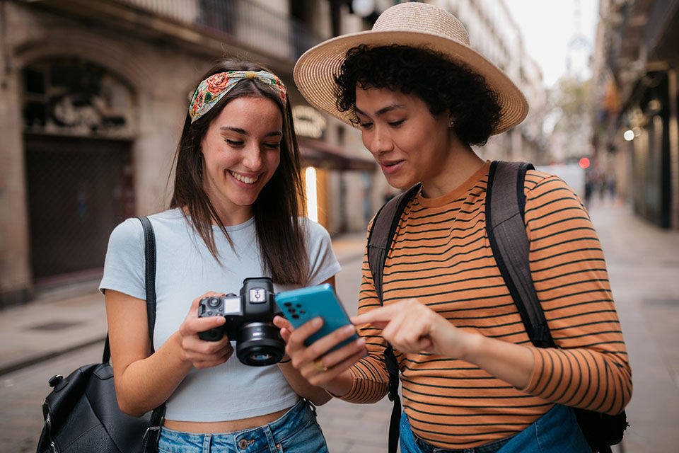 Happy diverse romantic couple of female tourists walking the streets of Barcelona together and looking at the photographs they took with their camera and a smartphone.