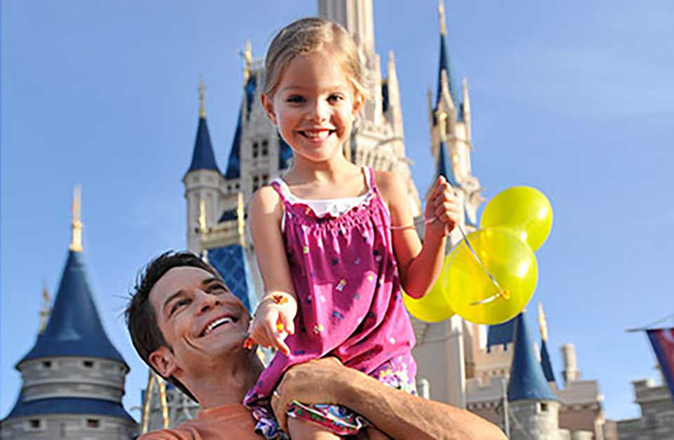 Father has young daughter on top of his shoulders why they both smile and she holds onto her bright yellow balloons.