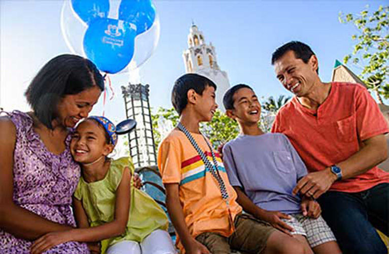 Family walks through disneyland holding a mickey mouse balloon smiling .