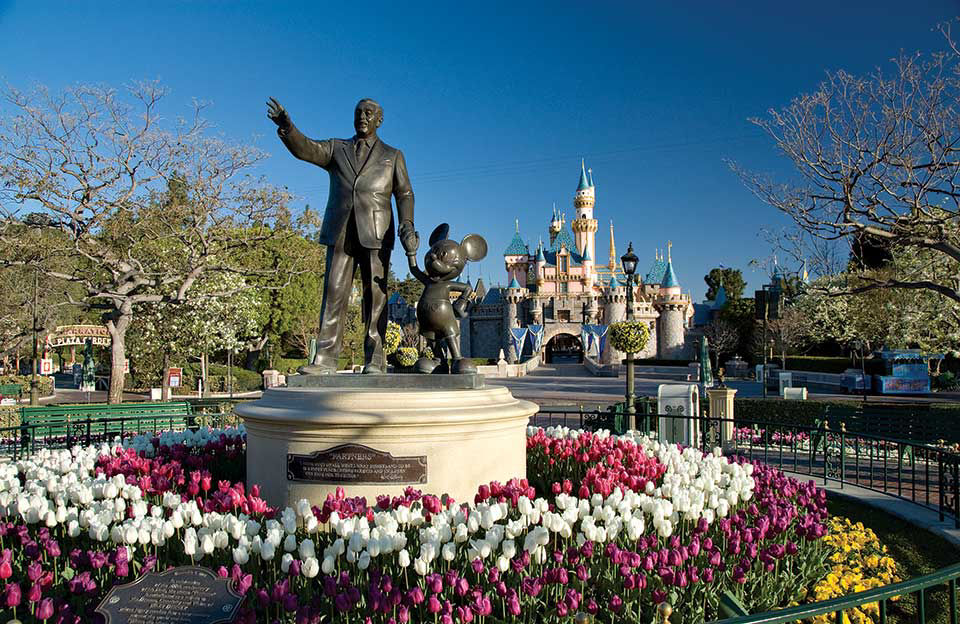 Statue of founder of disney holding mickey mouse hand surrounded by flowers with a castle backdrop.