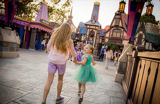 Daughter in bright green tulle dress excitedly grabs mothers hand as they walk into the park.