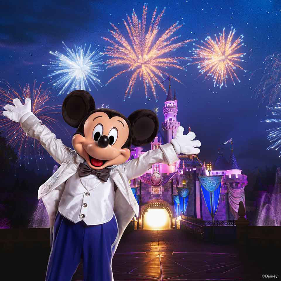 Mickey mouse celebrates with his hands spread out why fireworks go off in front of the castle at disneyland.
