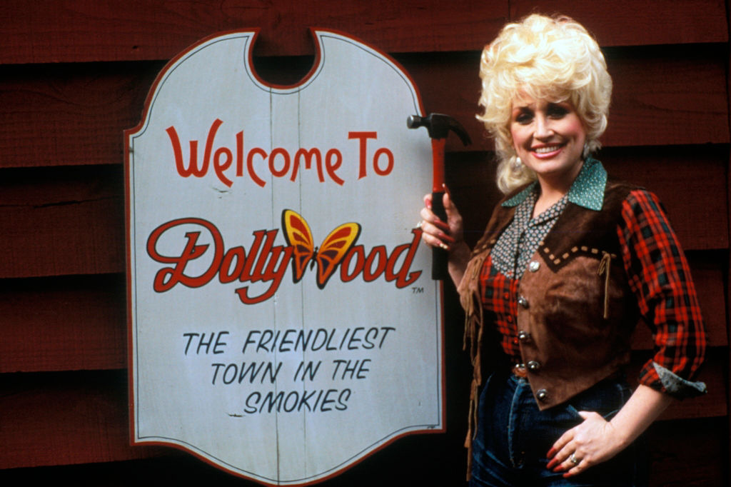 A young Dolly Parton stands next to the Dollywood sign.