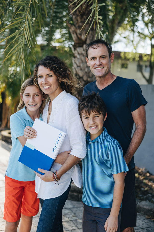 A smiling family of four stands outdoors under palm trees, with the mother holding a blue and white Estate Plan binder.