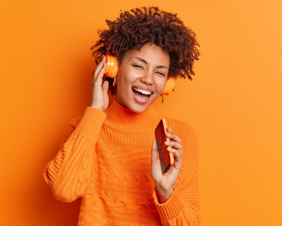 Woman in an orange sweater enjoying music through her headphones while holding her phone.