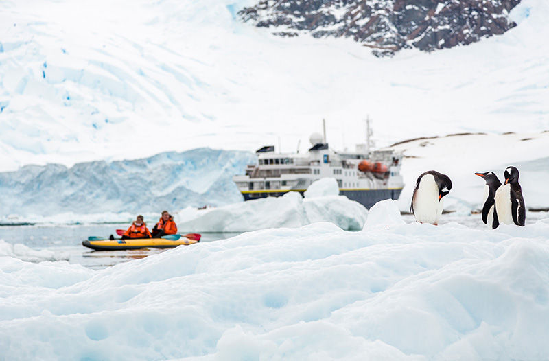 An expedition ship docked off an icy island with penquins in the foreground and a group of tourists in a small boat off the shore.